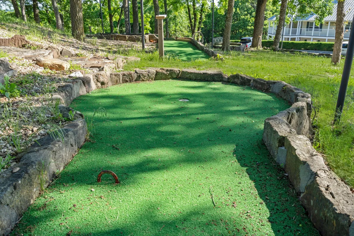 A playful miniature golf course nestled among mature trees and bordered by stone, creating a fun and relaxed outdoor environment. The scene is inviting for family gatherings or friendly competitions, highlighting another layer of recreational opportunity.
