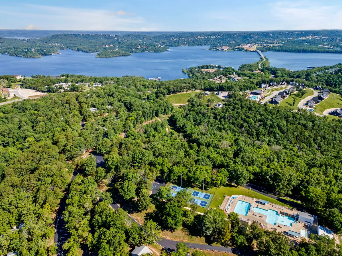 A panoramic view reveals an expansive lake bordered by lush green trees, swimming pools, tennis courts, and neighborhood homes, illustrating the full scope of outdoor activities available. The image captures the essence of lakeside living and the proximity of resort amenities to natural attractions.