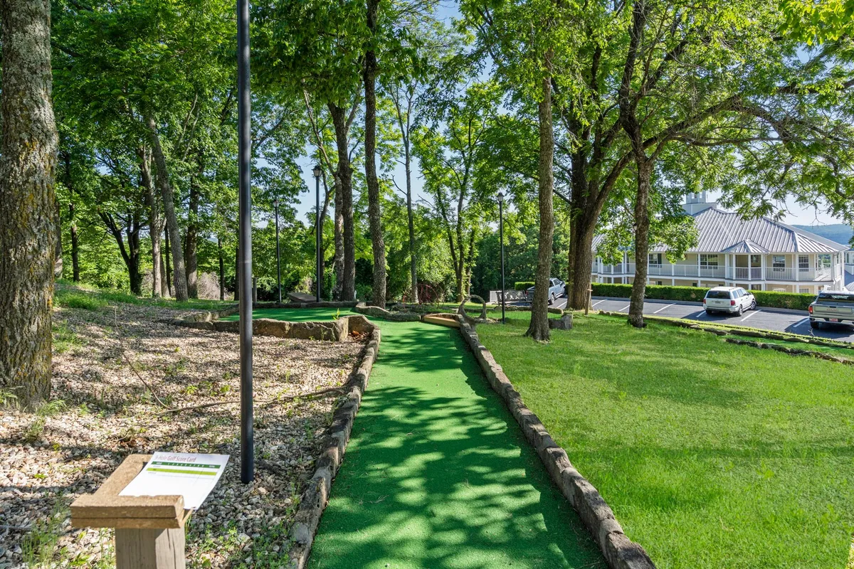 A mini-golf path winds through lush, green grass and is surrounded by mature trees. The expansive grassy area evokes a playful, relaxing mood and offers plenty of room for pets to roam while their owners enjoy leisure activities.