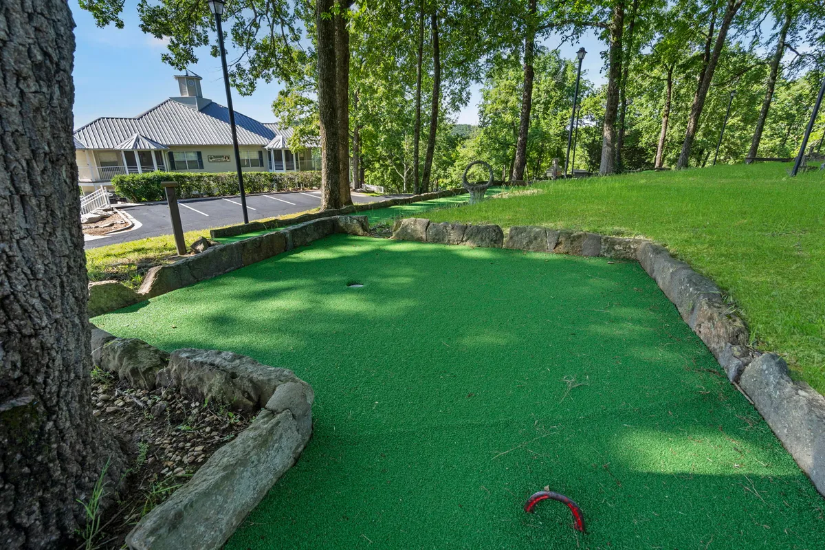 A playful and vibrant mini-golf area surrounded by lush green grass and tall trees, with stone boundaries defining the course. The setup welcomes families and children to engage in cheerful rounds of mini-golf, picnics, and outdoor games, creating an inviting and relaxed atmosphere.