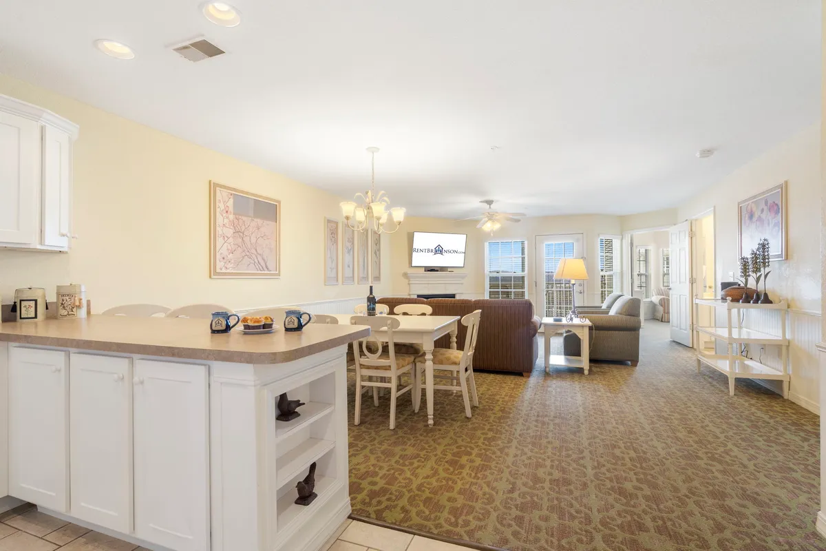 A sunlit open-concept living space featuring a plush sofa, a dining table set for four, a modern kitchen island with stools, stainless steel appliances, and decorative light fixtures. Large windows invite in natural light, creating a welcoming atmosphere that connects the kitchen, dining, and living areas seamlessly.