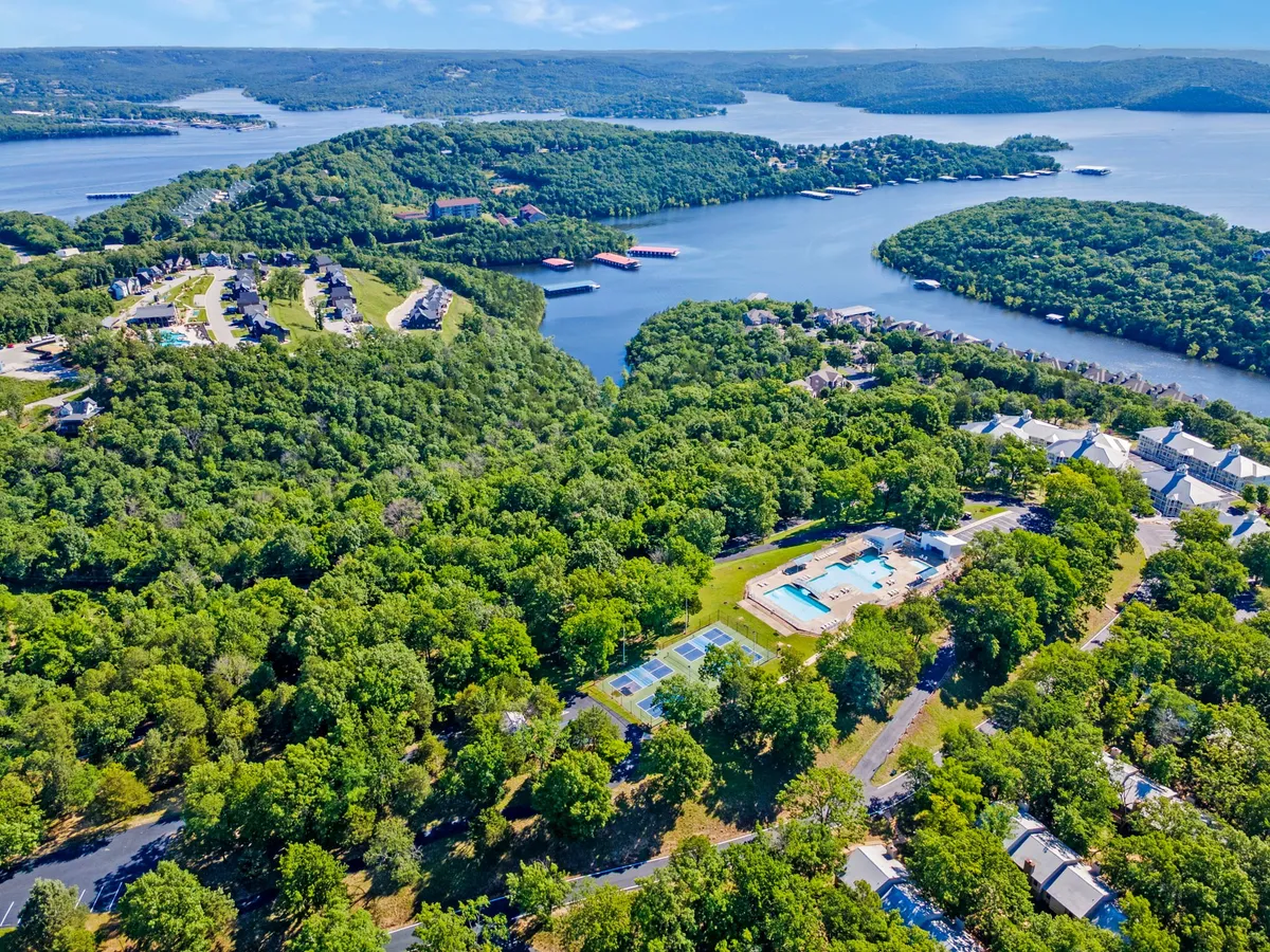 An active community scene featuring a sparkling swimming pool, tennis courts, lush greenery, and a waterfront backdrop with residential buildings. The image captures multiple amenities, including spaces for swimming, tennis, boating, and hiking, and conveys a lively, recreational atmosphere.
