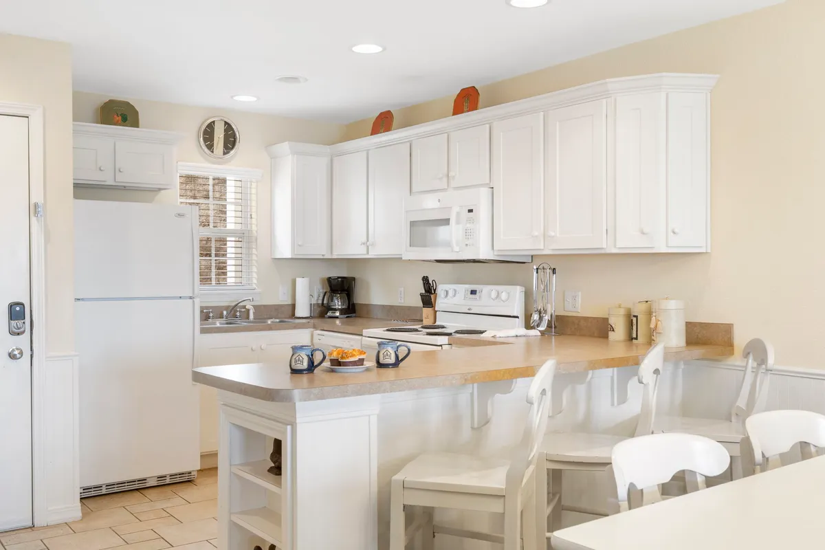A modern kitchen with a central island, crisp white cabinetry, ample counter space, and breakfast mugs arranged invitingly. The space radiates warmth and is set up for both cooking and casual gatherings, making it ideal for family meals or morning coffee.