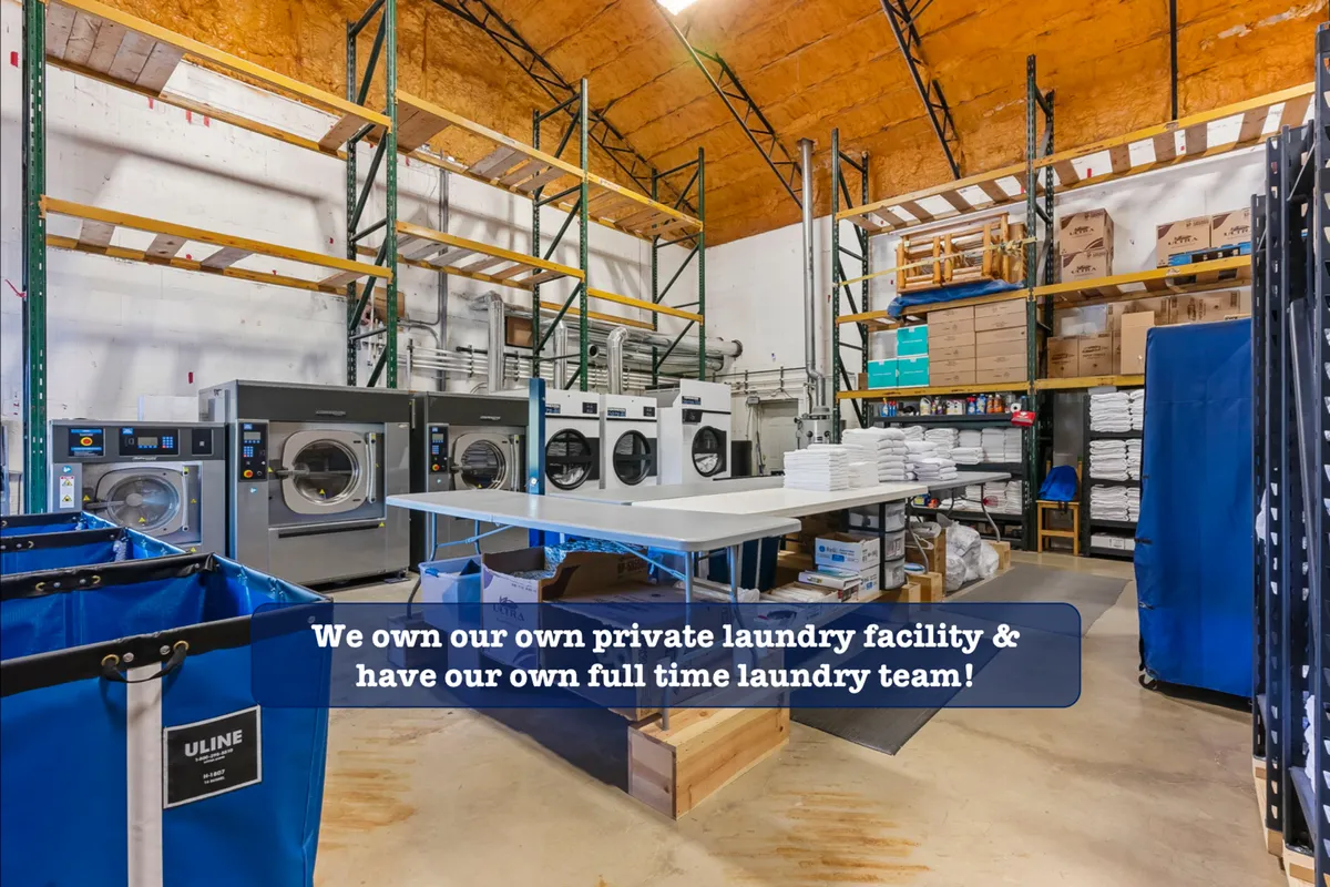 A professional-grade laundry room featuring industrial washers and dryers, organized storage shelves, and laundry bins. The space is brightly lit and immaculately clean, emphasizing efficient handling and processing of linens and towels.