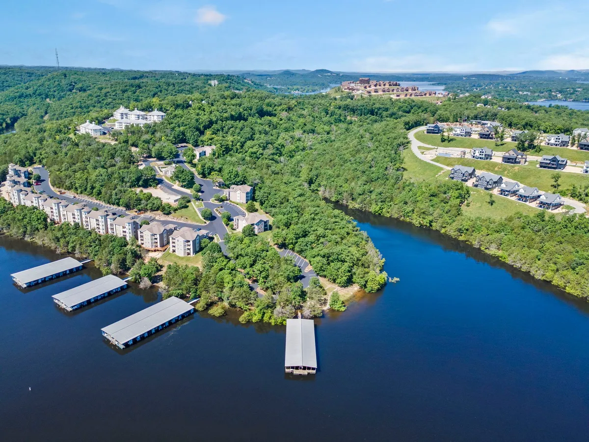 The image displays a tranquil lake bordered by lush greenery, with boathouses and residential buildings nestled along the shoreline. The scene suggests opportunities for lakeshore walks and relaxation by the water, all under the soothing ambiance of the Ozark Mountains.