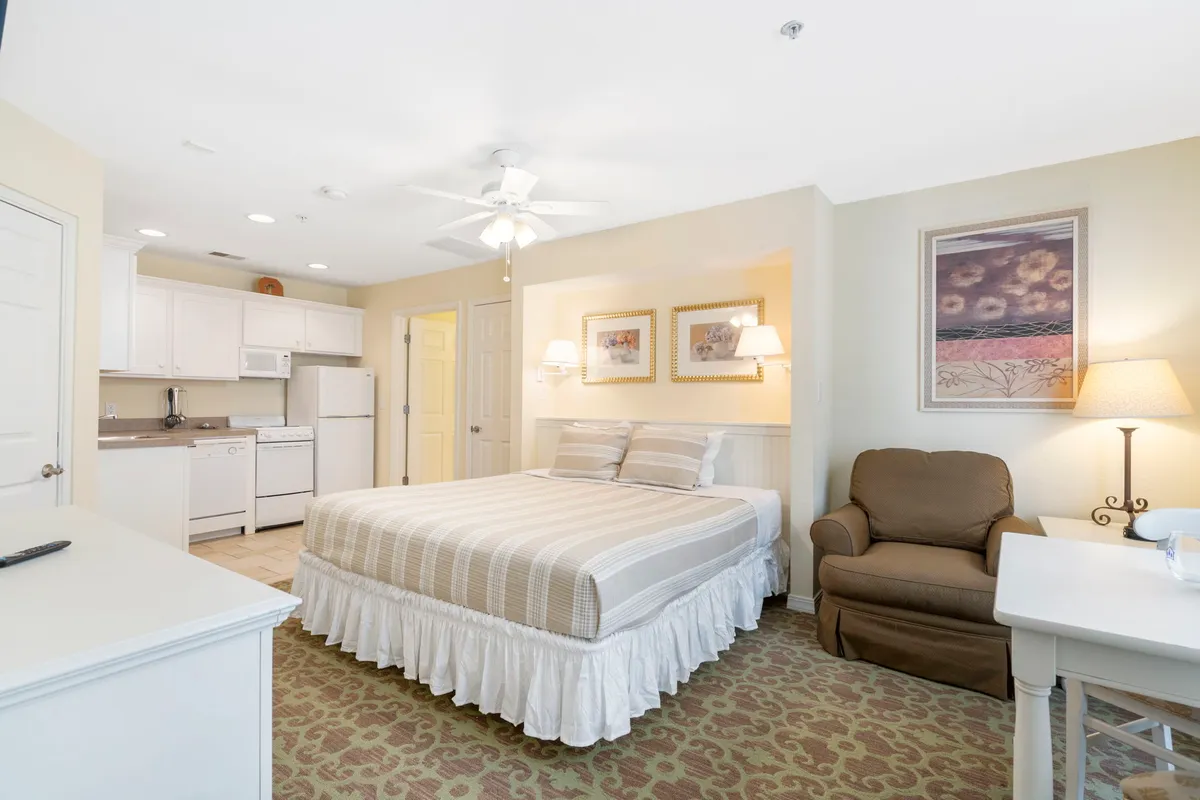 A welcoming bedroom featuring a cozy king bed, soft neutral bedding, an inviting armchair for reading, a compact kitchenette with appliances, and tasteful artwork on the walls. Sunlight filters into the space, enhancing its warmth and comfort.